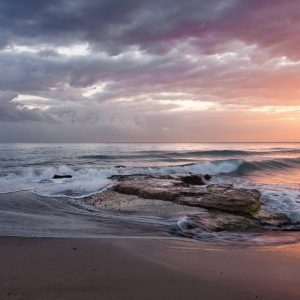 Sunset over a beach with a wave washing over a rock.